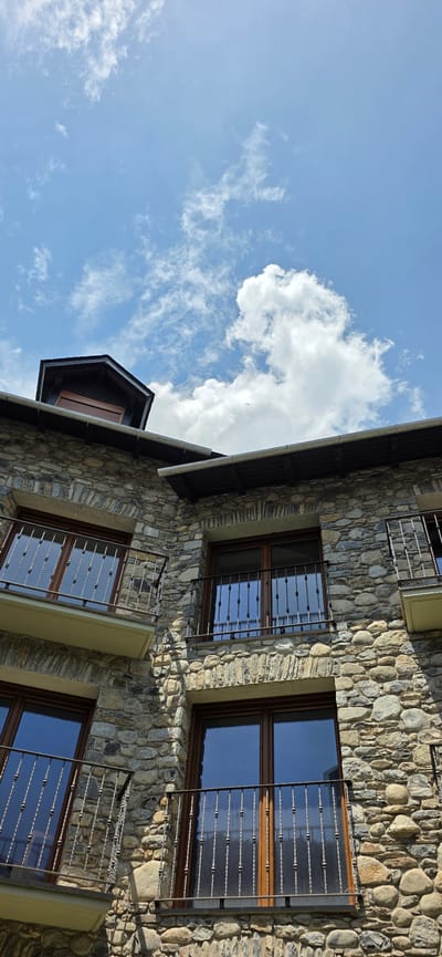 The image presents an upward view of a stone building set against a bright blue sky with scattered white clouds. The building's exterior is constructed of varying sizes and colors of stones. It includes windows with dark wood frames and decorative wrought iron bars, reflecting the sky. The perspective is angled, tilting the building diagonally across the frame. This image was probably taken in the city of Benasque, Spain.