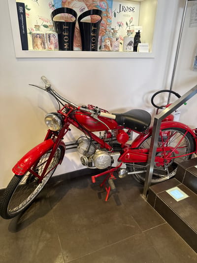 The image depicts a vintage red motorcycle parked indoors, leaning against a white wall. Above the motorcycle, on a shelf, are several decorative items, including bottles with artwork, two black Moët & Chandon ice buckets, and a framed picture. To the right of the motorcycle, a metal railing extends upwards, and a fire extinguisher is visible. The floor is dark brown tile. The scene appears to be inside a shop or establishment, likely in San Giovanni, Italy, given the context of the motorcycle and the decorative items suggesting a themed retail space. There are no people visible in the image.