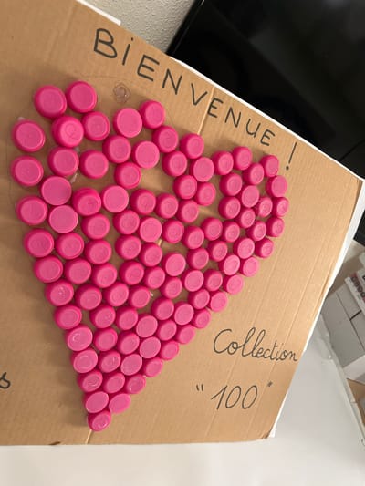 A slightly high-angle indoor shot depicts a brown cardboard box with handwriting and pink bottle caps arranged in a heart shape. The words "BIENVENUE!" are written on the left side of the box. On the upper right, "Collection" and "100" are written. The pink bottle caps, each with a white logo, form a heart shape, glued to the cardboard. A flat-screen television can be partially seen in the upper left corner, while what appears to be a white desk or shelf is on the right. The location is the city of La Fouillouse, France.