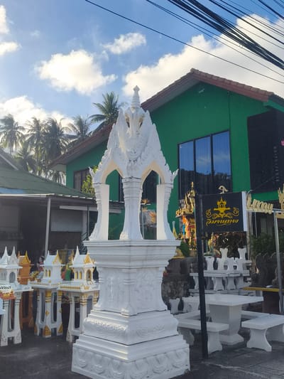 A collection of small, ornate spirit houses and a large white shrine are displayed outdoors in front of a bright green building. The spirit houses are intricately designed, many with white walls and gold accents, featuring tiered roofs and decorative pillars. The large central shrine is also white, with elaborate carvings and a multi-tiered roof structure. In the background, the green building has large windows reflecting the sky and palm trees. A sign with yellow Thai script on a black background is visible. The scene appears to be set in a shop or marketplace specializing in these religious structures, likely in Baan Thong Po, Thailand. Palm trees are visible, suggesting a tropical location. The sky is blue with scattered white clouds, and the lighting indicates it is daytime, possibly morning or afternoon. There are no people clearly visible in the image, but the offerings and decorations suggest a place of worship or commerce related to spiritual practices.