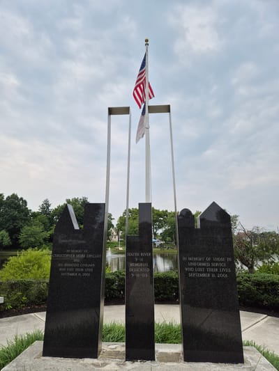 This image depicts a 9/11 memorial in South River, United States of America. The memorial consists of several polished black granite slabs, some of which are shaped with angled tops and curves. An American flag waves on a flagpole in the foreground.

One of the granite slabs is inscribed with "IN MEMORY OF CHRISTOPHER MORE DINCUFF AND ALL INNOCENT CIVILIANS WHO LOST THEIR LIVES SEPTEMBER 11, 2001." Another slab reads "SOUTH RIVER 9-11 MEMORIAL DEDICATED 9-11-03." A third slab is inscribed with "IN MEMORY OF THOSE IN UNIFORMED SERVICE WHO LOST THEIR LIVES SEPTEMBER 11, 2001."

Behind the memorial, a body of water can be seen, possibly a pond or lake, with a fountain. Lush green trees and bushes form the background, and a few buildings are visible in the distance. The sky is overcast with some clouds. The overall mood conveyed by the memorial and its setting is somber and respectful, commemorating the lives lost on September 11, 2001. The time of day appears to be daytime, given the lighting.
