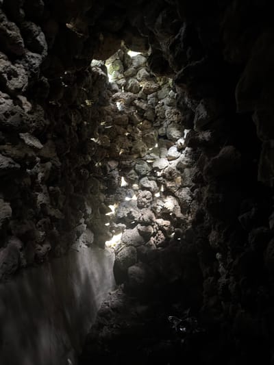 The image captures the interior of a dark, cavernous space, likely a cave or grotto, characterized by rough, dark grey rock formations. The scene is dominated by natural light filtering through numerous irregular openings between the rocks, primarily on the left and central parts of the frame. These bright shafts of light create dramatic contrasts, illuminating the rugged, porous texture of the stone walls and casting deep shadows in other areas.
The ceiling and walls are composed of densely packed, irregular stones, possibly volcanic rock, which create a raw and untamed atmosphere. No people are visible, and no specific activity is taking place. On the right side of the frame, a smoother, lighter-toned pathway or floor is discernible, suggesting a maintained area for visitors to traverse the dark environment. This pathway contrasts with the wilder, more chaotic rock formations. The strong directional light from outside indicates it is daytime. The overall impression is one of natural beauty and geological interest, with the interplay of light and shadow highlighting the cave's unique features. No text is visible in the image.