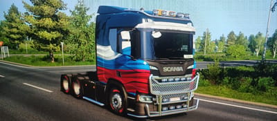 An image displays a custom-painted Scania truck with a Russian flag design on its side, driving on a road. The truck is dark blue on top, with white and red stripes below, mimicking the Russian flag. The truck has several lights on its roof and a large chrome grille with additional lights. A license plate is visible on the front bumper, reading "CR 833 PI". The truck appears to be in motion, as indicated by the slight blur on the tires and the road. The surroundings are green and lush, with trees lining the road, suggesting a rural or suburban setting. The sky is clear and blue. The image is a screenshot, likely from a video game, given the graphical style and the user interface elements that might be present but are not clearly visible. The location is stated to be Mokhsogollokh, Russia, which is a village in the Republic of Sakha (Yakutia), known for its natural landscapes.