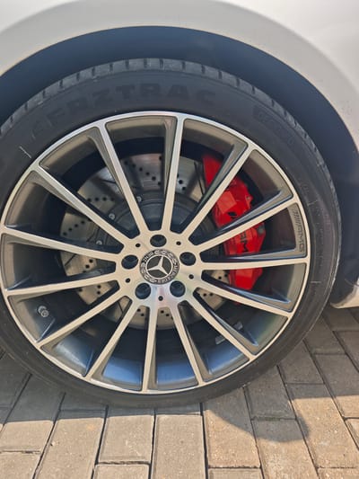 The image shows a close-up of a Mercedes-Benz AMG wheel with red brake calipers. The tire has text that appears to say "FRZTRAC DESIGNED IN GERMANY". The car is parked on a paved surface, possibly in an outdoor setting like a driveway or street. The background is blurred, but it appears to be a sunny day. There are no people visible in the image. The car is white.
