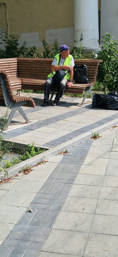 A man wearing a high-visibility vest and a blue cap sits on a wooden park bench, holding a cigarette. He has a dark bag slung over his shoulder and a black plastic bag on the bench next to him. Another black plastic bag lies on the ground to the right of the bench. The bench is made of horizontal wooden slats and metal supports. The ground is paved with light and dark grey rectangular tiles, with a concrete curb and some vegetation bordering the tiled area. In the background, there is a pale yellow wall with some white patches and large white columns. The scene appears to be outdoors, in a public space, possibly a park or pedestrian area. The man is facing to the right, looking away from the camera. The lighting suggests it is daytime, likely sunny. The image does not contain any explicit visual cues that definitively place it in Miass, Russia.