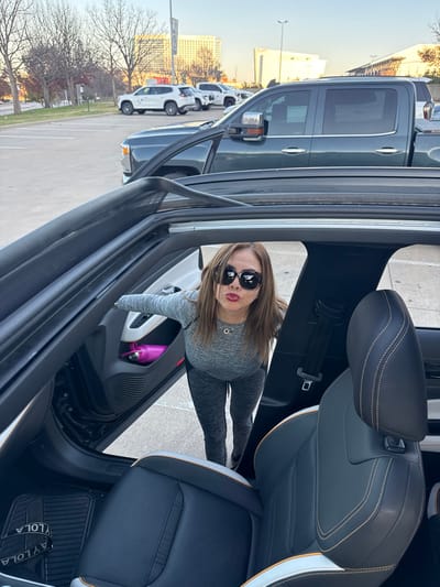 A woman in sunglasses and gray workout clothes poses in a car. The car door is open, and a pink water bottle is visible in the door's storage area. The interior features black leather seats with white and orange accents. Outside, a parking lot contains several trucks. A building is visible in the distance. The image is rotated counter-clockwise.