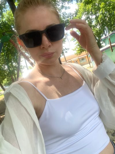 A fair-skinned woman with blonde hair pulled back, wearing black sunglasses and a white tank top with a sheer white cover-up, poses for a selfie outdoors. She is outdoors with trees and sunlight visible in the background, suggesting a park or a yard. The woman is adjusting her sunglasses with her right hand. The overall tone of the image is casual and summery.