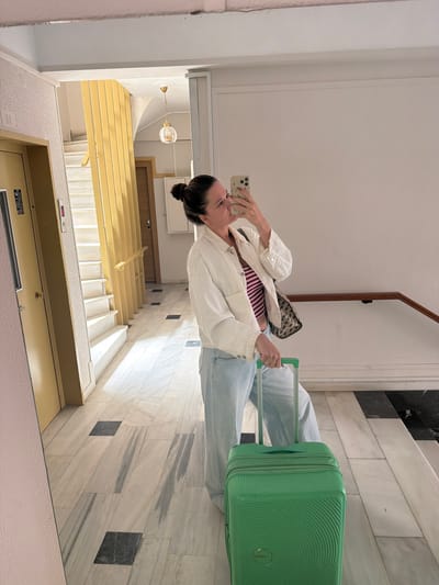A woman is taking a selfie in what appears to be a mirrored hallway or lobby in Glifada, Greece. She is wearing a white jacket, jeans, and a striped top, and is holding a green suitcase. The hallway features marble walls and floors, an elevator, and a staircase leading down. A light fixture hangs in the top corner of the image.
