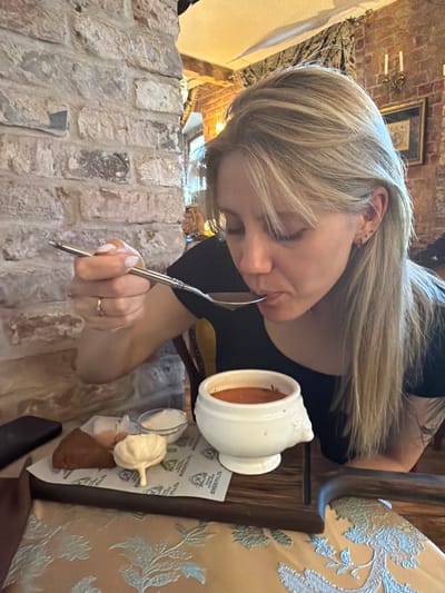 In Suzdal, Russia, a woman with blonde hair is seated at a table in a restaurant with a brick wall interior. She is eating a bowl of soup with a spoon, bringing it to her mouth. On the table in front of her is a wooden board with a small bowl of soup, a small glass of white sauce, a piece of bread, and a decorative element resembling a garlic bulb. The table is covered with a light-colored tablecloth with a floral pattern. The background shows more of the brick wall and a framed picture.