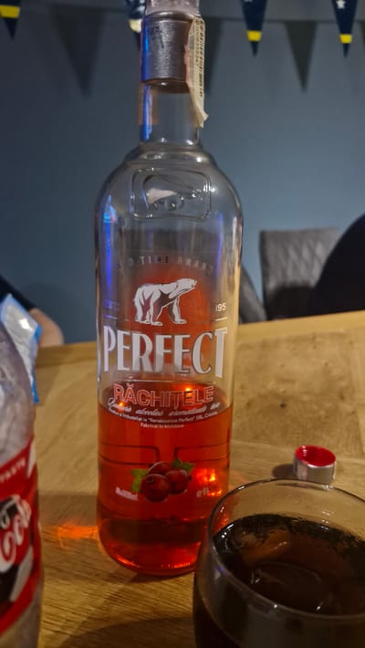 A high-angle, close-up shot shows a bottle of Perfect Răchițele flavored alcoholic drink, placed on a wooden table. The bottle is mostly transparent, revealing a reddish liquid inside. A polar bear logo is visible on the label, along with the text "PERFECT RĂCHIȚELE". Other elements in the scene include a partially visible bottle of Coca-Cola on the left, a glass filled with a dark beverage and ice on the right, and a red bottle cap lying nearby. In the background, there's a blurry depiction of a person and a gray chair, with some decorative flags hanging above. The lighting is dim, with blue and yellow hues present in the background. This scene is taking place in Uithuizen, Netherlands.