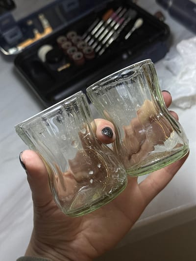 A close-up, slightly angled indoor shot shows a person's hand holding two small, clear glass bottles with a subtle light green tint. The bottles are rectangular in shape with fluted sides and appear to be stuck together or held very tightly. White, possibly dried, residue is visible on the interior and exterior surfaces of the glass. The person's thumb and index finger are visible, with dark, glittery nail polish on the fingernails.
In the blurred background to the left, a black case is open, revealing a collection of tools, likely makeup brushes and applicators, arranged neatly. Several small, circular, metallic objects, possibly rings or beads, are also indistinctly visible within the case. The lighting is bright, suggesting an indoor setting. There is no visible text in the image. The scene is set in Kuda, Russia.