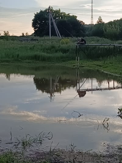 A person is fishing from a wooden platform over a body of water. The person is wearing a black shirt and is seated. A fishing rod is extended over the water. The scene is set in Verkhnie Chershily, Russia, with a background of trees and a radio tower. The water reflects the surrounding environment and the sky. The foreground shows the edge of the water with grass and some debris. The time appears to be late afternoon or early evening, given the lighting.