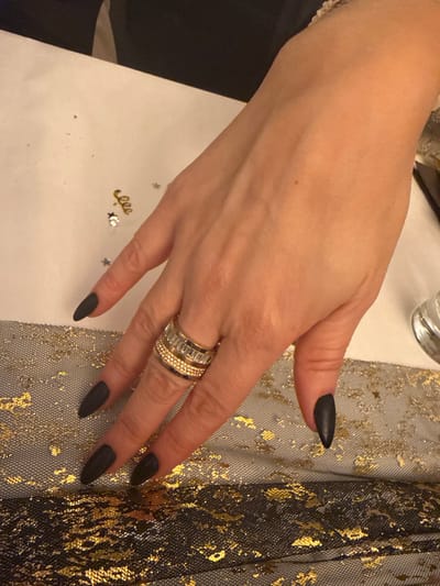 A hand adorns black, sharp, pointed nails and a stack of gold and diamond rings on the ring finger. The hand rests upon a surface decorated with gold stars, a "eee" embellishment, and a gold-splattered mesh fabric. The overall image suggests a festive or celebratory occasion.