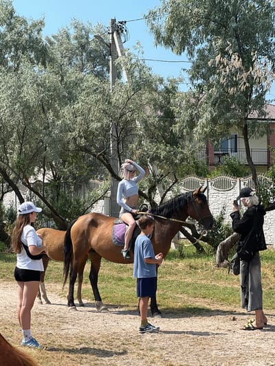 The image shows a sunny outdoor scene with several people and a horse. The setting appears to be a rural or semi-rural area, possibly a farm or stable, with trees and dry, dusty ground. A brown horse is the central focus, with a young woman in a light blue outfit riding it. She has blonde hair and glasses. Another person, a young man in a blue t-shirt and dark shorts, stands beside the horse, appearing to assist or guide it. Further back, an older person in a black jacket and gray pants is looking at their phone and holding a black bag. Another young woman wearing a white t-shirt and black shorts, with a white cap, is sitting on the ground, looking towards the horse. The overall impression is of a casual, daytime activity, likely horseback riding or training, in a warm weather environment. The location cues suggest it is in Mysivske rural settlement, Ukraine. No visible text is present in the image.