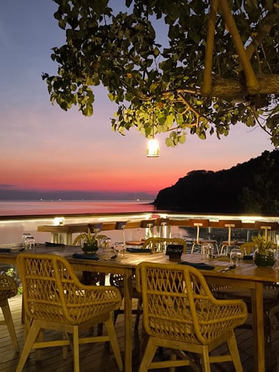 The image captures a serene waterfront restaurant setting in Labuan Bajo, Indonesia, during twilight. The foreground is dominated by a wooden table set for dining, complete with glassware, plates, and napkins. Woven rattan chairs with wooden legs surround the table. A small vase containing greenery sits on the table. In the background, the calm sea stretches out to a silhouetted hill. The sky displays a gradient of colors from deep blue at the top to vibrant orange and pink near the horizon, indicating sunset. A single lantern hangs from a tree branch above the dining area, casting a warm glow. The overall ambiance is tranquil and inviting, suggestive of a peaceful evening meal by the coast.
