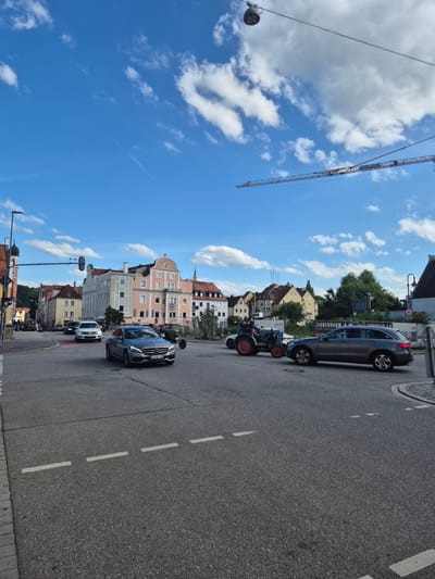 This image shows a street scene in Landshut, Germany, with a focus on an intersection. In the foreground, there is asphalt road with white lane markings. A grey Mercedes-Benz sedan is driving in the right lane. To the left of the Mercedes, a blue tractor with two people on it is also in the intersection, seemingly stopped or moving slowly. Behind the tractor, a dark grey SUV is in the right lane, also in the intersection.
In the background, there are several buildings, predominantly in pastel colors, with a mix of architectural styles. Some of the buildings appear to be older, with tiled roofs. A construction crane is visible in the sky, indicating ongoing construction in the area. The sky is blue with scattered white clouds. Traffic lights and street lamps are also present. The scene appears to be from a daytime setting, with clear weather conditions.