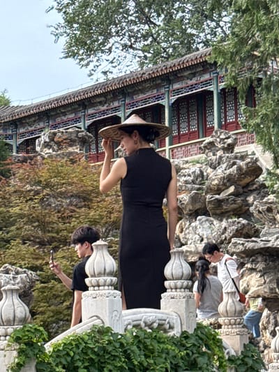 In the Xicheng District, China, at what appears to be a traditional Chinese garden or park, a woman in a black dress and a conical straw hat stands on a white stone balustrade, adjusting her hat. She is the primary subject in the foreground. To her left, a young man in a black t-shirt is captured in profile, holding a smartphone and appearing to take a photo. Behind and to the right of the woman, a man in a white shirt with glasses and a dark bag slung over his shoulder is visible, along with a woman in a grey t-shirt. Further to the right, another person in a green shirt and blue pants is partially visible. The background features a multi-tiered building with ornate red and blue decorative elements and a tiled roof, as well as rocky landscaping and lush green trees. The overall scene is one of people visiting a cultural landmark.