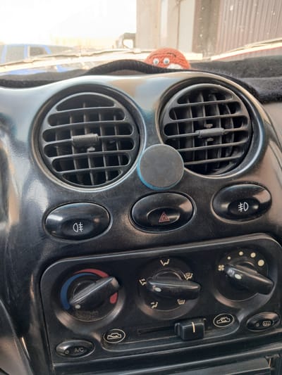 The image shows a close-up view of a car's dashboard. The dashboard is predominantly black and features several controls and air vents. In the center, there are two round air vents positioned side-by-side. Below the vents, there are several buttons and knobs for controlling the car's climate and lighting. One button displays a fog light symbol, another a hazard light symbol, and a third a front fog light symbol. There are also rotary knobs for temperature and fan speed, as well as a slider control. An "A/C" button is visible on the lower left. A small, crocheted orange and white bobble with googly eyes sits on the dashboard above the right air vent. The dashboard appears slightly dusty. The background suggests an outdoor setting, possibly a parking area, with a blurred view of other vehicles and structures. The image composition focuses on the interior of the car.