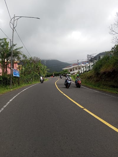The image captures a curved asphalt road through a mountainous and vegetated area in Saing Ta Mawt Lay, Myanmar, under an overcast grey sky. The road is marked with a solid white line on its left edge and a solid yellow center line, indicating a leftward curve from the viewer's perspective. Several motorbikes are in motion. In the mid-ground, three riders on motorbikes proceed towards the viewer: one on a dark scooter with a light-colored helmet, another on a dark scooter with a green helmet and green apparel, and a third on a red scooter with a green helmet and dark jacket. Additional motorbikes are visible further down the curved road. On the left, a tall grey street light pole with a curved arm and LED fixture stands beside dense green foliage, including palm trees. Partially visible behind the vegetation is a pink-colored building and a blue banner. On the right, a steep, green, vegetated embankment borders the road, with a white multi-story building featuring a blue-tiled roof and complex structure visible on the hillside above. Dark green mountains, partially obscured by cloud cover or fog, dominate the distant background. Power lines extend across the scene.