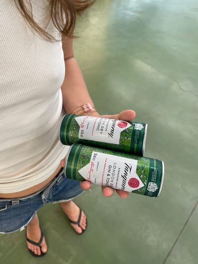 A person wearing a white ribbed tank top and denim shorts is holding two cans of Tanqueray London Dry Gin & Tonic in their right hand. The cans have a green and white design with floral patterns. The person is standing on a green, smooth surface that appears to be a floor. The scene takes place indoors, possibly in a store or warehouse, given the lighting and floor surface. The image does not contain enough information to determine the specific location within Pizarra, Spain.