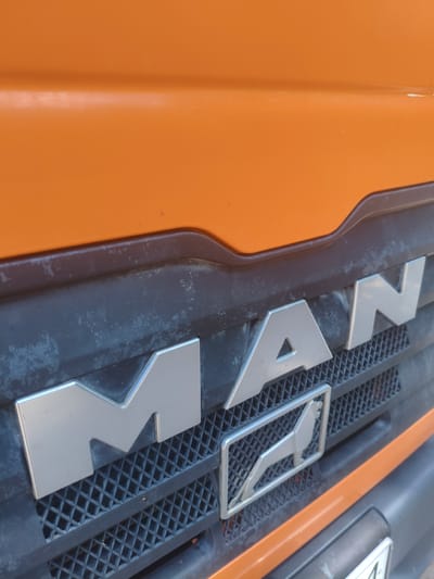 The image shows a close-up view of the front grille of a MAN truck. The grille is black and has a diamond pattern. The letters "M", "A", and "N" are prominently displayed in silver chrome on the grille. Below the letters, there is a silver emblem of a lion. The truck's body, which is orange, is visible at the top of the image. The scene appears to be outdoors. The location is identified as Alchevsk, Ukraine.