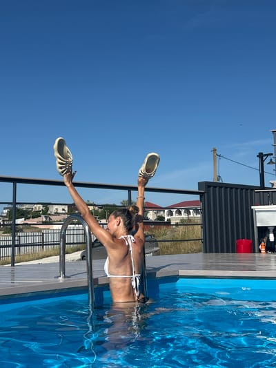 A woman in a white bikini is standing in a bright blue swimming pool, holding a pair of cream-colored slides above her head. She is positioned near the pool ladder, with her back to the camera and her arms raised. The water is clear and reflects sunlight. The pool is surrounded by a wooden deck and a metal railing. In the background, there are buildings with red roofs, a fence, and a clear blue sky. The scene appears to be outdoors, likely in a residential area. The location is identified as Mysivske settlement, Ukraine.