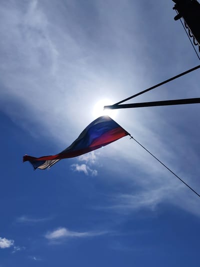 The image shows the flag of Russia flying in front of a bright sun, likely in Kosino, Russia. The sky is blue with some wispy clouds. The flag is tattered at the edges. There are dark, metal poles visible in the upper right corner of the frame, suggesting a flagpole structure.