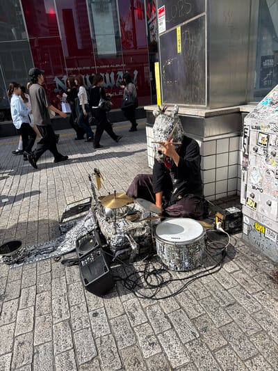 The image depicts a street scene in Shibuya, Japan, with a pedestrian walkway paved with grey cobblestones. In the foreground, a street performer is seated on the ground, wearing a distinctive mask resembling a stylized animal head with visible hair. The performer is playing a drum set, which includes at least one snare drum with the "EVANS" logo, a cymbal, and a bass drum adorned with a patterned wrap. Two black speakers are positioned near the drum set, connected by cables. A dark purple cloth is draped over the performer's lap.
Behind the performer, a group of people are walking along the sidewalk. Some are looking at their phones, while others appear to be engaged in conversation. The background features a building with a large glass facade that reflects the surrounding city. A sign on the building reads "TREASURE 4th MINI ALBUM NEW". To the right, a metal structure, possibly part of a subway entrance or kiosk, is covered in various stickers and graffiti, including what appears to be a "STAR WARS POP-UP STORE" advertisement. The overall impression is of a busy urban environment with diverse elements of street culture and commerce.