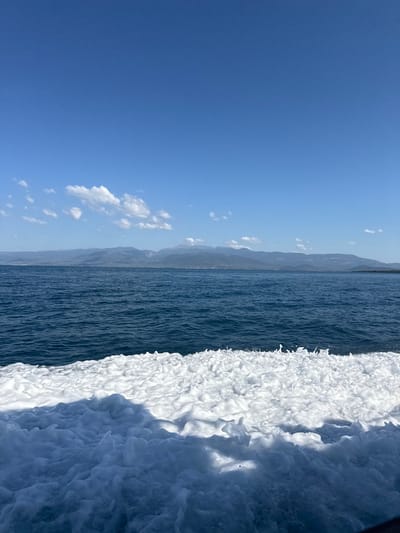The image shows a view of the sea from a boat, with white foam churned up by the boat in the foreground. The water is a deep blue, and beyond it, a distant coastline is visible with mountains in the background. The sky is a clear blue with a few white clouds. The scene appears to be located on the waters off the coast of Stroggili, Greece, based on the provided context. There are no people or specific objects clearly identifiable in the image other than the natural elements of the sea, sky, and land. The action depicted is the movement of the water and the resulting foam. The environment is a marine landscape under a clear sky.