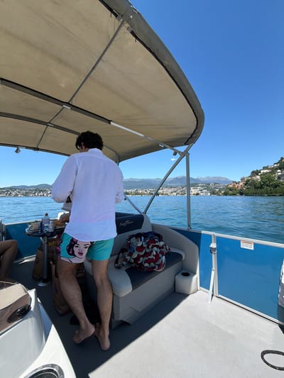 A man wearing a white linen shirt and teal swim trunks with a cartoon image on them stands on a pontoon boat on Lake Lugano in Switzerland. He is facing away from the camera and appears to be preparing food on a small table. A bottle of water and other items are on the table. The boat has a tan canopy and blue railing. In the background, the city of Lugano is visible on the shore, with buildings and mountains in the distance. The water is blue and calm.
