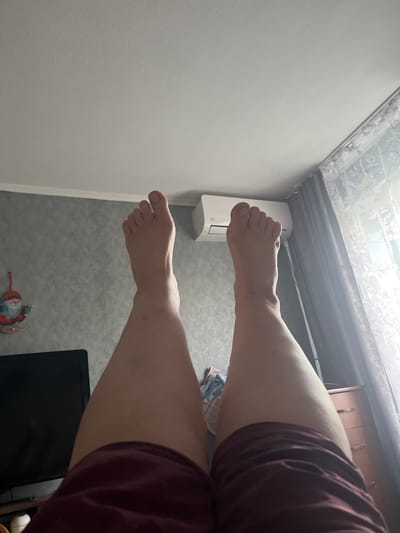 The image shows a person's legs and feet in a room. The person is wearing maroon shorts. Their feet are positioned in the foreground, with the toes of one foot clearly visible. In the background, there is a white air conditioning unit mounted on a grey wall. Above the air conditioner, a ceiling is visible with a patterned design and what appears to be a metal structure. To the right, there are wooden drawers or cabinets. The overall lighting suggests it is daytime, possibly indoors with artificial light. There are no visible text elements in the image. The scene is likely a residential setting.