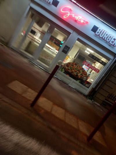 A dimly lit storefront with "Gusto Burger" signage is visible. The neon "Gusto" sign glows red. A pizza is displayed in the window along with a sign reading "UNE OFFRE". The storefront's entrance is a glass door, and part of the interior is visible. The sidewalk is dimly lit, and there are two red posts partially visible in the foreground. The image has a slightly blurred, tilted perspective.