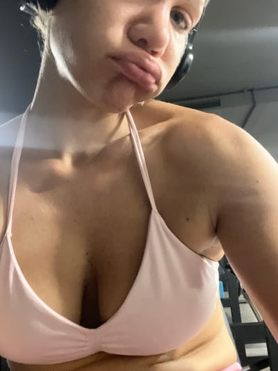 The image shows a close-up of a woman's upper body in what appears to be a gym. She is wearing a light pink bikini top and black headphones. Her face is turned slightly away from the camera, with her lips puckered. The background is out of focus and shows gym equipment. There is no identifiable information within the image to place it in Mühldorf a. Inn, Germany.