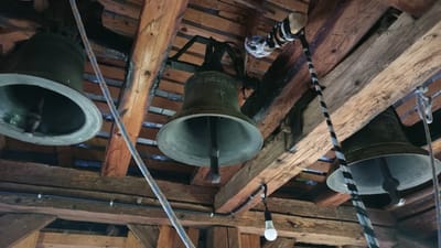 The image shows a close-up view of several large, dark-colored bells suspended from wooden beams in what appears to be a belfry. The bells are made of metal and have a classic bell shape, with clappers visible inside. Ropes are attached to the bells, suggesting they are meant to be rung. The wooden structure is rustic and appears old, with visible grain and some charring on certain beams, possibly from age or past events.
In the background, a single white light bulb is illuminated, attached to a wire that runs along the beams. There are also electrical cables and conduits visible, indicating that the belfry has been equipped with modern lighting. The overall scene is dimly lit, with light primarily coming from the single bulb and some ambient light filtering from above or through unseen openings. The perspective is from below, looking upwards at the bells and the wooden ceiling of the belfry.
The scene is static, with no human activity directly depicted, though the presence of ropes implies past or potential use for ringing the bells. The focus is on the bells themselves and the structural elements of the belfry. The setting suggests a historical or religious location, likely a church or bell tower, given the presence of bells. The condition of the wood and the rustic nature of the structure point towards an older building. No text is visible in the image. The location context provided is Vyshka, Ukraine.