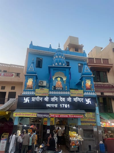 The image shows the exterior of a vibrant blue temple in Amritsar, India, identified by signage as the "Prachin Shri Shani Dev Ji Mandir" (Ancient Lord Shani Dev Temple). The temple features ornate blue architecture with tiered roofs and decorative finials, adorned with images of deities within small alcoves. A prominent black sign with white lettering reads "प्राचीन श्री शनि देव जी मन्दिर" (Prachin Shri Shani Dev Ji Mandir) and "स्थापना 1761" (Established 1761). Yellow banners with text in Hindi and Punjabi are displayed, one of which announces "विशाल भण्डारा 30 जून 2026 दिन मंगलवार" (Grand Feast June 30, 2026, Tuesday).
In front of the temple entrance, which is partially obscured by a metal gate and various offerings, a crowd of people are visible. Some are dressed in traditional Indian attire, including women in kurtis and scarves, and men in shirts and t-shirts. Several individuals are gathered around a stall selling religious items and food. A motorcycle is parked near the entrance, and other buildings with shops and residential elements can be seen flanking the temple. The overall scene depicts a busy street in Amritsar, likely during daytime, with religious activity and commerce coexisting.