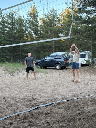 Two adult males are playing volleyball on a sandy court surrounded by pine trees. One male, wearing a gray t-shirt and black shorts, stands on the left side of the court, facing right. The other male, wearing a tan t-shirt and blue shorts, is in mid-air on the right side of the court, with his arms raised in a spiking motion. A volleyball is visible above the net between them. In the background, a dark colored SUV and a white camper trailer are parked. The scene appears to be in a recreational area near Ozerki (Krasnodolinskaya), Russia, given the pine forest environment.