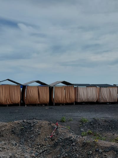 The image depicts a row of closed beach cabanas with brown and beige curtains, set against a backdrop of a cloudy sky and a dark sandy beach. The cabanas have peaked roofs and appear to be constructed from metal frames. In the foreground, a pile of rocks and gravel is visible, with some sparse green vegetation growing amongst them. A red and white striped rope or barrier is tied to a pole in the rock pile. The scene is devoid of people. The location is specified as the city of Ақши, Kazakhstan.