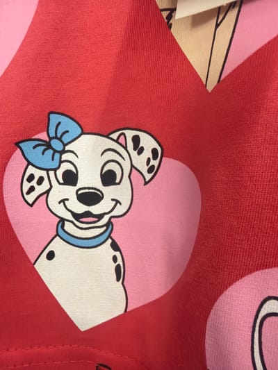 The image is a close-up, high-angle shot of a red fabric with a cartoon Dalmatian dog printed on it. The dog is white with black spots and is wearing a blue bow tie and a blue collar. It is depicted within a light pink heart shape. The fabric itself has a textured appearance. Parts of other shapes, possibly other hearts or parts of a pattern, are visible in pink and red. A thin white strip of material and some black lines are visible in the upper left quadrant of the image, suggesting the edge of another fabric or garment. There is no visible text in the image. The lighting suggests an indoor setting. The overall impression is of a children's clothing item or fabric.