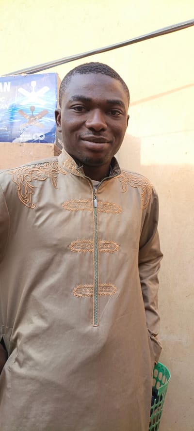 A man poses indoors, wearing a tan, ornate, zippered jacket. The jacket features elaborate embroidery around the shoulders and down the zipper. A box with a ceiling fan illustration sits slightly behind him. His expression is soft, with a slight smile. A green plastic basket is visible at the bottom right of the frame. The background appears to be a plain, light-colored wall.