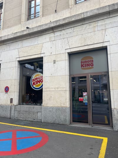 A daytime view captures the street-level facade of a Burger King restaurant in Genève, Switzerland. The building features a light-colored, possibly stone or marble, exterior with horizontal banding and a darker granite base.
The main entrance consists of glass doors framed in metal, situated to the right. To the left of the doors, a large window displays the modern Burger King "bun" logo, which is orange and white with "BURGER KING" text, partially visible through a frosted or tinted section of the glass. Below this, a smaller window showcases the older, circular Burger King logo with a blue circle, red "BURGER KING" text, and yellow accents, hanging inside the clear glass.
The asphalt street occupies the lower and right portions of the frame. A solid yellow line is painted on the asphalt, running parallel to the building. Prominently displayed on the asphalt is a large, circular red and blue symbol, indicating a "no parking" or "no stopping" zone, featuring a red border, a blue interior, and a red diagonal line. A smaller, identical "no parking/stopping" sign, a red circle with a blue background and a diagonal red line, is affixed to the building wall just below the lower Burger King window.
The lighting suggests it is daytime, likely overcast, as there are no harsh shadows. Reflections of other buildings and the sky are visible in the glass surfaces of the windows and doors. No people are present, and no specific activity is taking place. The only clearly readable text is "BURGER KING" on both logos.