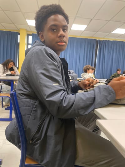 The image portrays an indoor classroom or lecture hall located in Hérouville-Saint-Clair, France. The central subject is a young Black male, appearing to be a teenager, seated at a light-colored desk in a blue metal chair with a wooden seat. He wears a dark grey or blue padded jacket over dark clothing and light grey pants. His dark, curly hair is styled high. He directs a neutral, steady gaze towards the viewer, with his lips slightly parted. His left hand rests on the desk, holding a thin black cord or strap, while his right forearm and hand are partially visible, also resting on the desk surface.
In the mid-ground to the left, a young female with long, dark hair and a white long-sleeved top is seated at a separate light-colored desk, engaged with a mobile phone held in her hands. A yellow structural pillar is visible behind her, adjacent to a window section. Further in the background, to the right, multiple other individuals, mostly young people, are seated at similar desks; one wears a grey hoodie, another a dark green top. The environment features light-colored walls, a ceiling with white rectangular acoustic tiles and recessed fluorescent light fixtures, and prominent blue curtains covering a wall section. The scene is evenly illuminated by artificial overhead lighting.