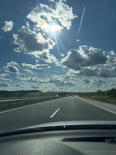The image presents a view from inside a vehicle traveling on a highway near Zlatina, Bulgaria. To the left is a bright, sunlit sky filled with cumulus clouds and a contrail from an aircraft. The vehicle is on a paved road with lane markings and adjacent grassy areas leading to a distant treeline or hills. The interior of the car is visible on the right.
