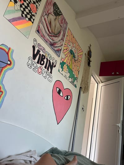 The image shows a bedroom with several pieces of art displayed on a white wall. One piece is a painting of a woman's back, with her hair tied up. Another painting features a psychedelic mushroom design with a radiating sun and flowers. There is also a graphic art piece with a checkered pattern and rainbow stripes. Below the paintings, the word "VIBIN'" is written in bold, stylized letters, surrounded by smiling face emojis. A large pink heart with cartoon eyes is also painted on the wall. The room appears to be in a domestic setting, with a bed visible in the foreground and a doorway in the background. The scene is brightly lit. There are no people clearly visible, other than a portion of a person's leg and foot on the bed. The location is identified as being in the city of Bulgarovo, Bulgaria.