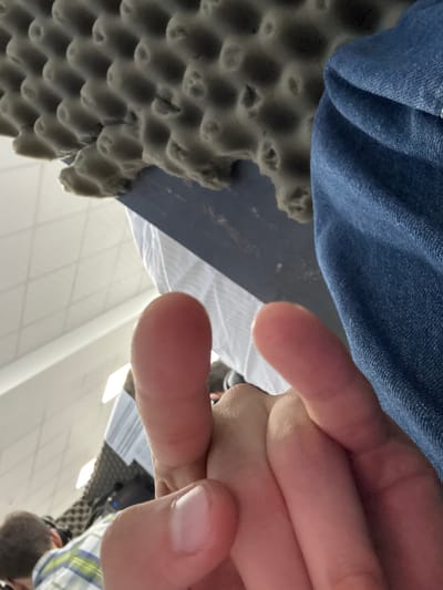 This is a close-up, low-angle shot of a person's hand with two fingers extended, appearing to point upwards. The fingers are pale pink and show natural skin texture. Behind the hand, to the left, is a textured, grey material, possibly acoustic foam, arranged in a grid pattern. Below the foam, a dark blue surface is visible, possibly a table or desk. To the right of the foam and blue surface, a portion of blue denim fabric, likely from jeans or a jacket, is present. The background is blurred but suggests an indoor environment with white tiled flooring and fluorescent lighting. In the far background, a person wearing a plaid shirt is partially visible, along with a black object that may be electronic equipment. The image is taken from a very low perspective, looking upwards towards the hand and the objects above. The specific location of Katta Chilanzar-2 MFY, Uzbekistan cannot be determined from the visual information present in the image.