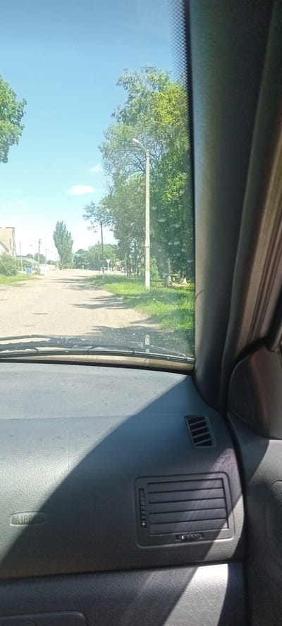 The image captures a view from the inside of a vehicle, looking through the windshield. The scene depicts a rural road lined with trees and grass under a clear blue sky. A lamppost stands on the right side of the road, and in the distance, there are buildings and more trees. The dashboard of the car is visible in the foreground, with an "AIRBAG" marking on the left side and air vents on the right. The image appears to be taken during daytime, with sunlight casting shadows on the dashboard. The location is identified as Koviahy, Ukraine.