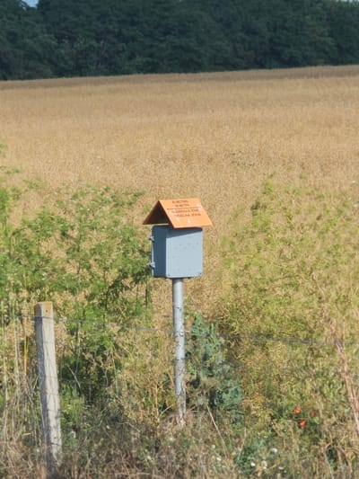 In Kolarovo, Bulgaria, a grey metal box with an orange, pitched roof sits atop a pole in a dry, grassy field. The box has a small, square flap and appears to be a weather station or monitoring device. Text on the orange roof reads "40 METERS 40 METRA HAZARDOUS ZONE ОПАСНА ЗОНА." A wire fence runs across the foreground, with a concrete post on the left. Beyond the field, a dense line of dark green trees forms the horizon. The sky is a pale blue.