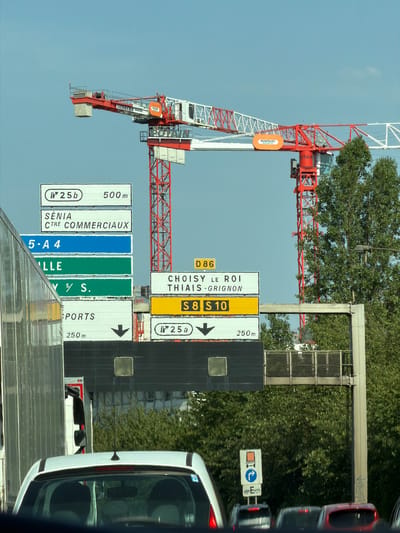 This is a rotated image of a street scene in Thiais, France, featuring a large red and white construction crane. The crane is the dominant object in the frame, with its boom extending horizontally across the middle of the image. Several road signs are visible beneath the crane, indicating directions and distances. One sign reads "CHOISY LE ROI THIAIS GRIGNON S8 S10" with an arrow pointing to the right and "250m". Another sign shows "500 m SÉNIA CTRE COMMERCIAUX 5-A4 LLE". There are also signs indicating "SPORTS 250m".
In the background, lush green trees are abundant, suggesting a summer day. The sky is clear and blue. A white truck is partially visible on the right side of the image, reflecting the surrounding environment. The overall impression is a bustling urban construction site with clear traffic signage. The text on the crane itself is partially visible, including "BOUYGUES" and "LIEBHERR". The image appears to have been taken during daylight hours. The orientation of the image suggests it may have been captured from a vehicle moving alongside the road. There are no people clearly visible in the image.