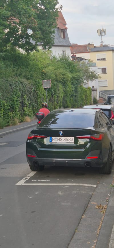 The image shows the rear of a dark green BMW parked on a street in Mainz, Germany. The car has a license plate that reads "M MS 3583". In the background, there are buildings and lush green trees. The sky appears to be overcast, suggesting a cloudy day. The street has a painted white line marking a parking space. A red object, possibly part of another vehicle or a street accessory, is visible behind the BMW. A square mirror with red and white stripes is mounted on a pole to the left of the car, possibly a traffic mirror. The scene appears to be a residential or urban street with a mix of architecture and nature. No people are clearly visible in the image.