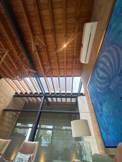 The image captures an interior architectural space in Valle de Bravo, Mexico. The ceiling features a blue mural reminiscent of a celestial scene with airships and geometric shapes. An air conditioning unit is mounted on the wooden paneled wall. Slatted wood elements run across the window, filtering light. Geometric, triangular light fixtures hang from the ceiling. Large glass panels reflect the scene, showing trees and outdoor elements, along with parts of furniture, including chairs with cushions. The room has a contemporary design with a blend of wooden and modern construction.