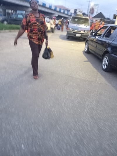 A woman walks along a paved road in Port-Harcourt, Nigeria, carrying a black and yellow bag in her right hand. She is wearing a multi-colored patterned shirt and dark leggings, with her left hand extended forward. A black sedan is parked on the right side of the road, and a light-colored van is in the background, also on the road. Other people and vehicles are visible in the distance, suggesting a busy urban environment. The sky is overcast.